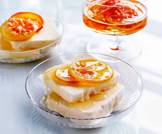 Slices of semifreddo topped with glazed orange slices on clear plates, with a glass of syrup in the background.