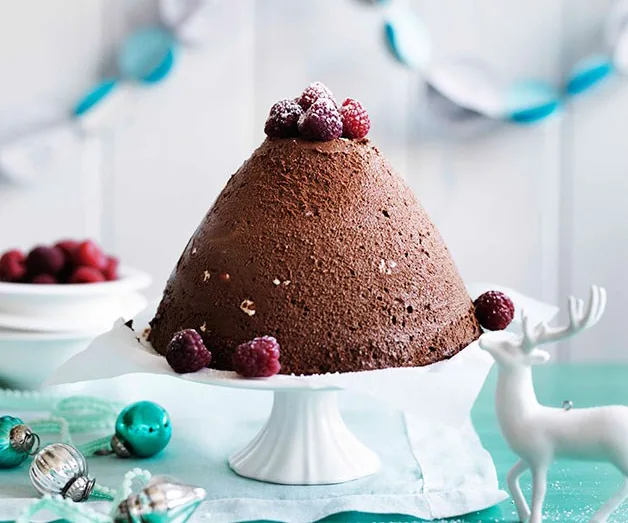 Chocolate dome cake with raspberries on top, displayed on a white cake stand, with festive decorations nearby.