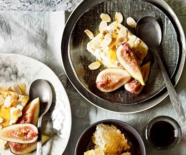 A dessert plate with figs, almonds, and honeycomb, accompanied by a spoon and a small cup of dark liquid.