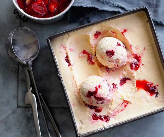 Ice cream with strawberry swirls and two scoops in a square pan beside an ice cream scoop and a bowl of strawberries.