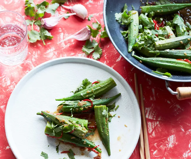 Plate and wok with stir-fried okra, garnished with cilantro and chili, on a red patterned table.