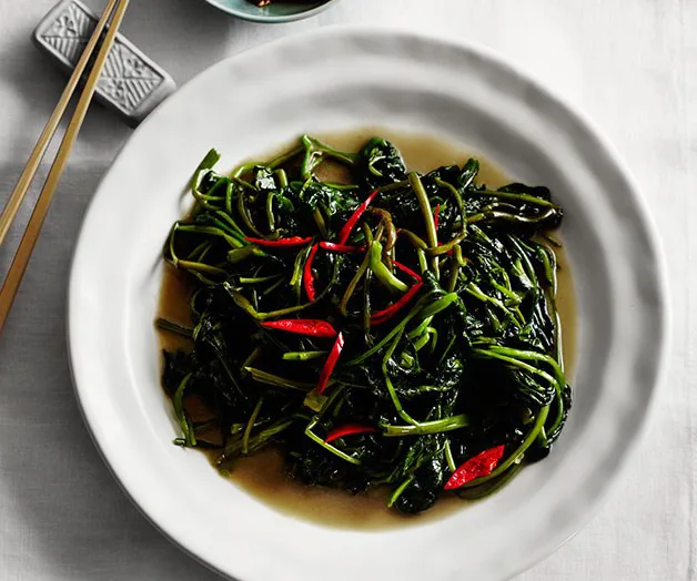 Stir-fried greens with red chilies on a white plate, accompanied by chopsticks on the side.