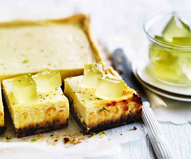 Lime cheesecake bars topped with lime jelly cubes on a baking sheet, with a dish of extra jelly cubes beside them.