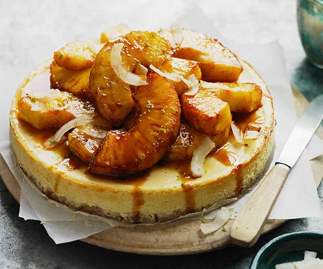 Caramelized pineapple cheesecake garnished with toasted coconut slices on a wooden board with a knife.