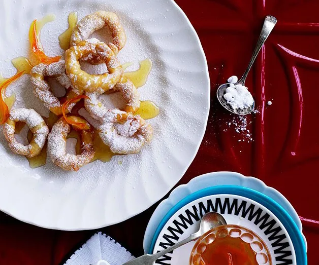 Plate of powdered sugar-dusted fritters with orange zest on a red table, beside a spoon of sugar.