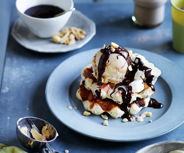 Waffles topped with ice cream, chocolate sauce, and nuts on a blue plate with a cup and spoon nearby.