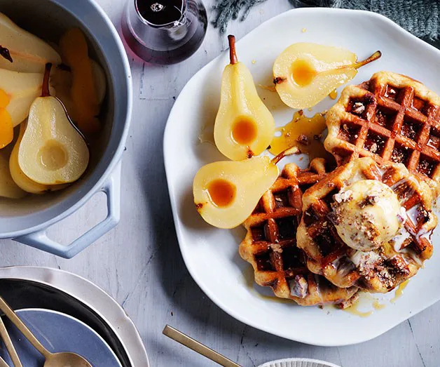 Waffles topped with ice cream, poached pears, and syrup on a white plate, with extra pears and syrup nearby.