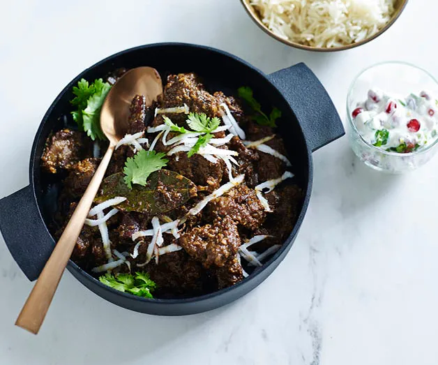 A pot of beef curry garnished with cilantro and shredded coconut, with rice and a yogurt dish on the side.