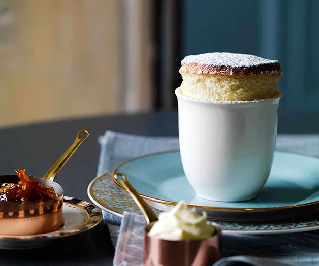 Plain white souffle with powdered sugar in a white cup on a saucer, with jam and cream in small copper dishes.
