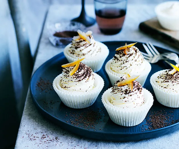 Creamy cupcakes topped with orange zest and chocolate shavings on a dark plate, with a glass in the background.