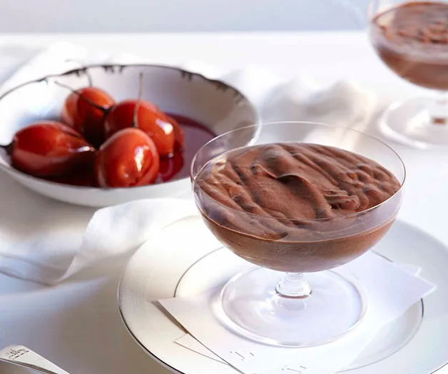 Chocolate mousse in a glass dish next to a bowl of poached pears in syrup on a white table setting.