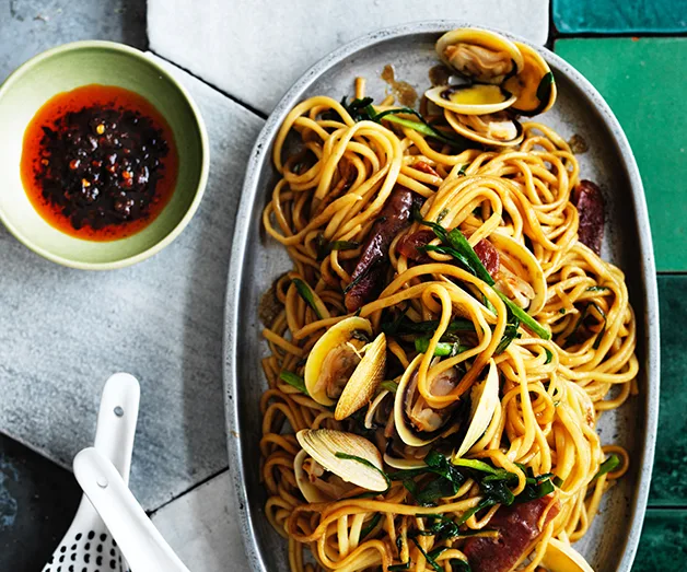 Plate of seafood noodles with clams and greens, accompanied by a small bowl of chili sauce.
