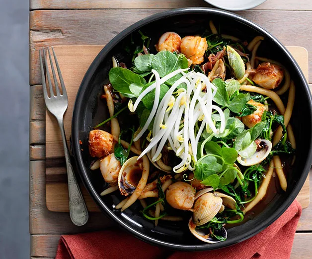 Seafood pasta with scallops, clams, greens, and bean sprouts on a black plate, fork beside it on a wooden table.