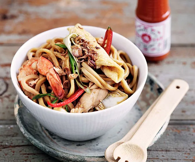 Bowl of noodles with shrimp, crab, and vegetables, garnished with herbs and accompanied by a bottle of hot sauce on a table.