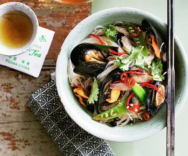 Bowl of seafood noodle soup with mussels, sliced chili, pea pods, and cilantro, next to a tea cup on a wooden table.