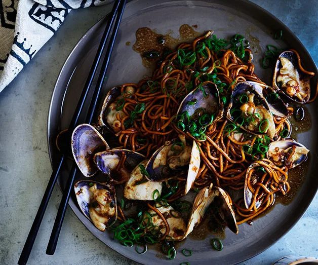Stir-fried noodles with clams and green onions served on a plate with chopsticks beside a cloth napkin.