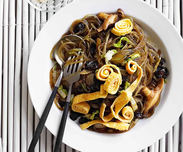 Stir-fried glass noodles with shiitake mushrooms and rolled omelet strips on a white plate with fork and chopsticks.