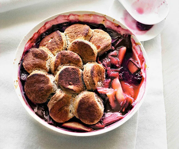 Bowl of rhubarb cobbler with golden biscuit topping, partially revealing the vibrant red fruit filling inside.