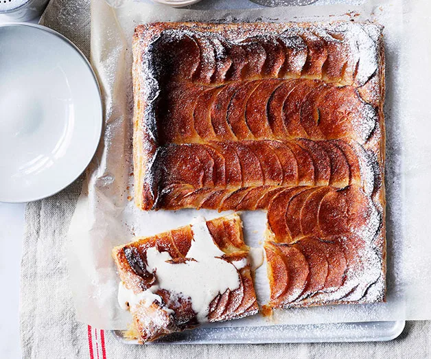 Rectangular apple tart with powdered sugar, one slice removed, placed on parchment, beside an empty white plate.
