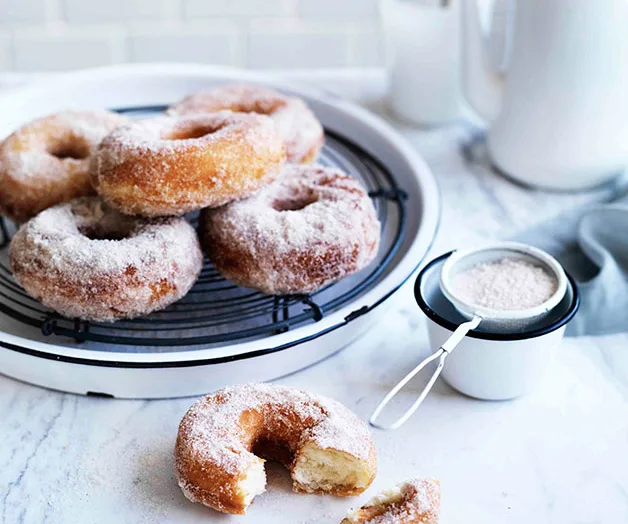 Sugar-coated doughnuts on a cooling rack, with one partially eaten, beside a cup of sugar on a marble countertop.