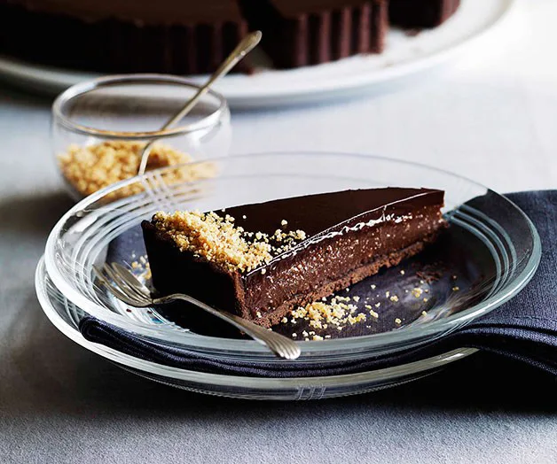 A slice of glossy chocolate tart topped with crumbled nuts on a clear plate, with a fork beside it.