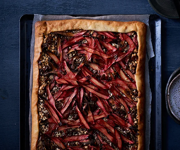 Rectangular tart with roasted pink rhubarb and walnuts on a dark baking tray.