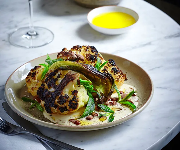 Grilled cauliflower with greens and sauce on a round plate, garnished with nuts, on a marble table with oil in a bowl.