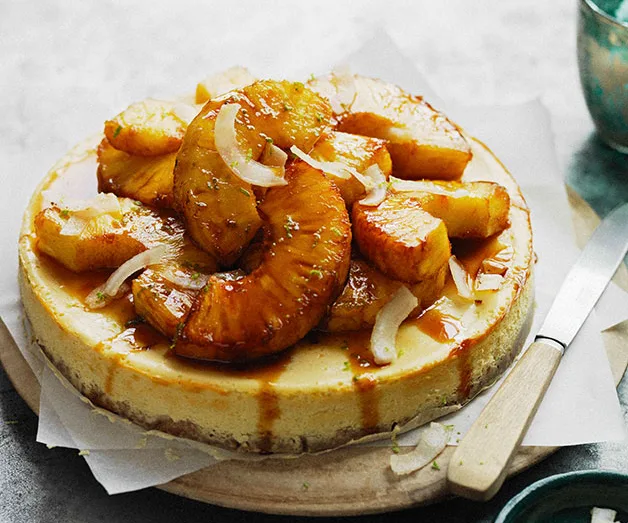Cheesecake topped with caramelized pineapple slices and coconut shavings on a wooden board, knife on the side.