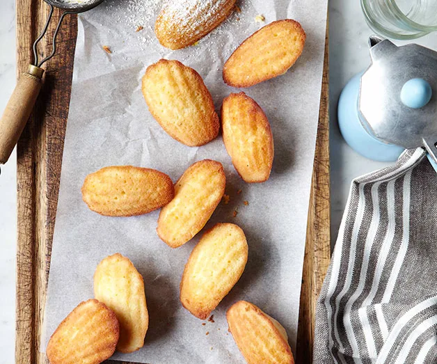 Browned madeleines on parchment paper with a dusting of powdered sugar, next to a striped cloth and pot.