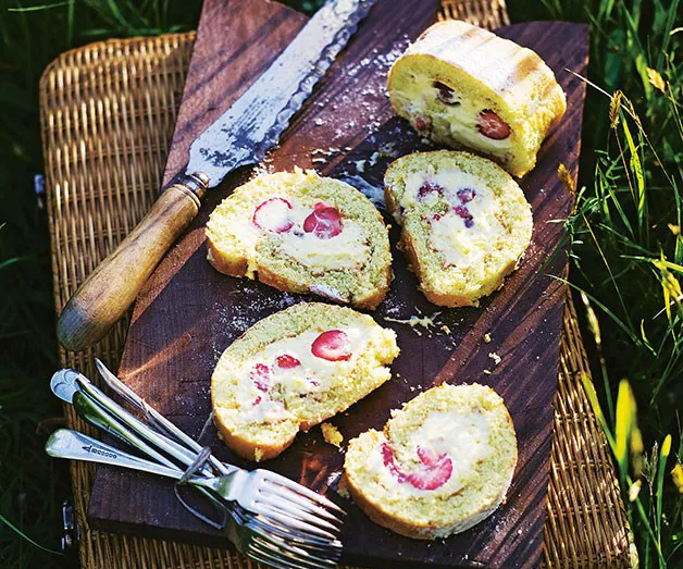 Sliced strawberry Swiss roll on a wooden board with a knife and forks beside on a picnic mat.