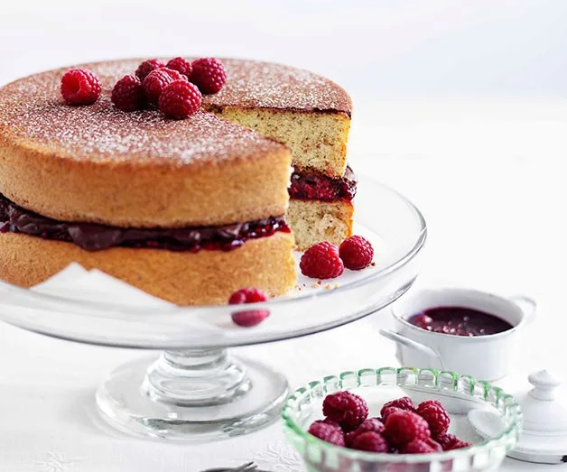 Sponge cake with raspberry filling and topped with fresh raspberries on a glass stand, next to raspberry sauce and bowl.