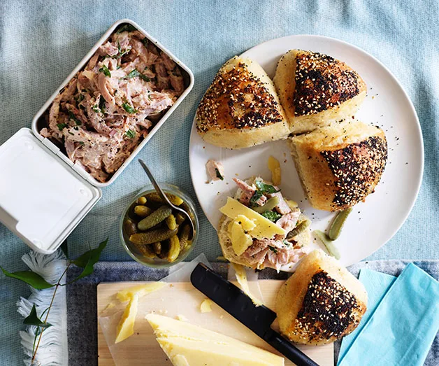 Bread rolls with pulled pork, cornichons, and cheese on a plate, with a tin of pork and a knife nearby on a blue cloth.