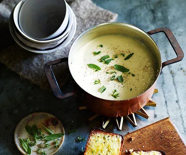 A copper pot of creamy soup garnished with fresh herbs, beside a plate of buttered bread and a stack of bowls.