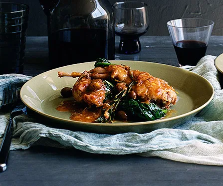 Braised rabbit with beans and greens on a plate, set on a table with a glass of red wine and a dark bottle.