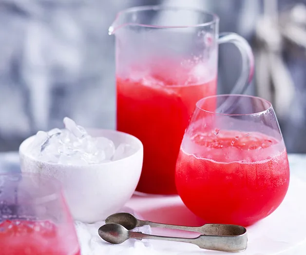 A glass and jug of red iced drink on table with two metal spoons and a bowl of ice.
