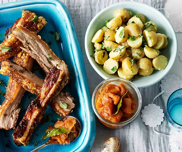 Baked ribs in a blue dish, with a bowl of seasoned potatoes and a jar of chutney on the side.