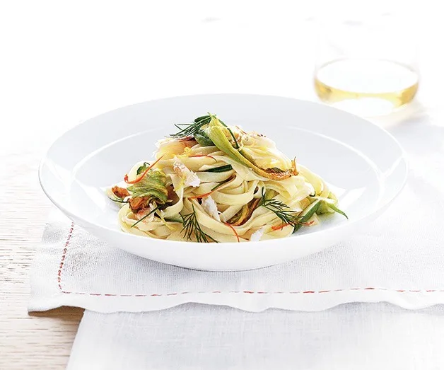 Fettuccine pasta with zucchini and herbs on a white plate, glass of white wine in the background.