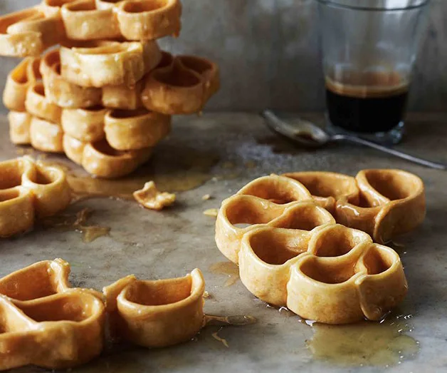 Flower-shaped waffles drizzled with syrup, stacked and scattered on a table, with a glass of coffee in the background.