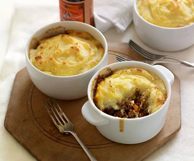 Three servings of shepherd's pie in white bowls on a wooden board with forks placed nearby.