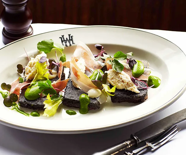 Plate of gourmet food with dark bread, sliced prosciutto, fresh greens, and vibrant green sauce, garnished elegantly.