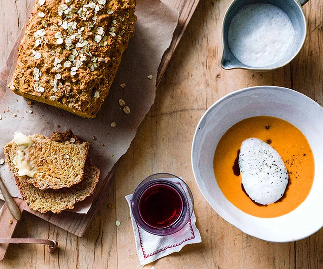 Loaf of oat bread with buttered slices, orange soup with cream, and a glass of red drink on a wooden table.