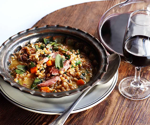 Stew with farro, carrots, and greens in a bowl, served with red wine on a wooden table.