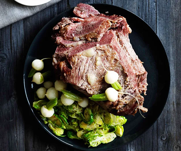 Corned beef with boiled baby potatoes, celery, and Brussels sprouts on a black plate on a wooden table.