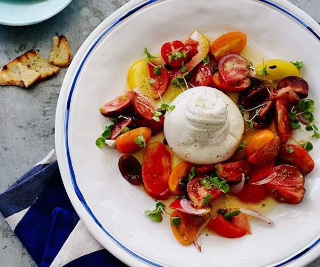 Fresh burrata salad with colorful cherry tomatoes and greens on a white plate, garnished with a drizzle of olive oil.