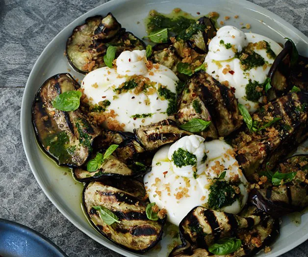 Grilled eggplant slices topped with creamy burrata, fresh basil, and herbs on a grey plate.