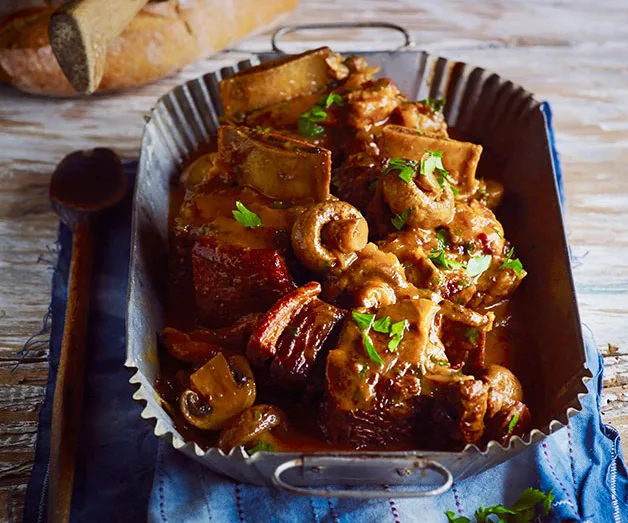Braised short ribs with mushrooms in a decorative dish, garnished with parsley, on a table with bread and spoon.
