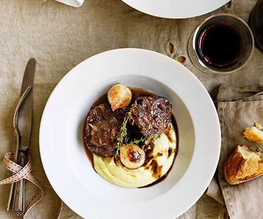 Braised beef with mashed potatoes and garnish on a white plate, served with a glass of red wine and sliced bread.