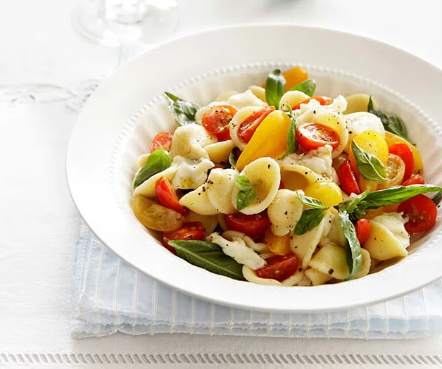 Colorful pasta salad with cherry tomatoes, mozzarella, and basil leaves in a white bowl on a light tablecloth.