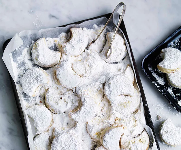 Tray of crescent-shaped, powdered sugar-coated cookies on baking paper with tongs.
