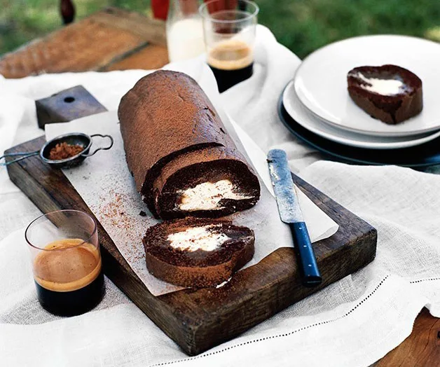 Chocolate roulade with cream filling sliced on a wooden board, surrounded by espresso, knife, and plates.